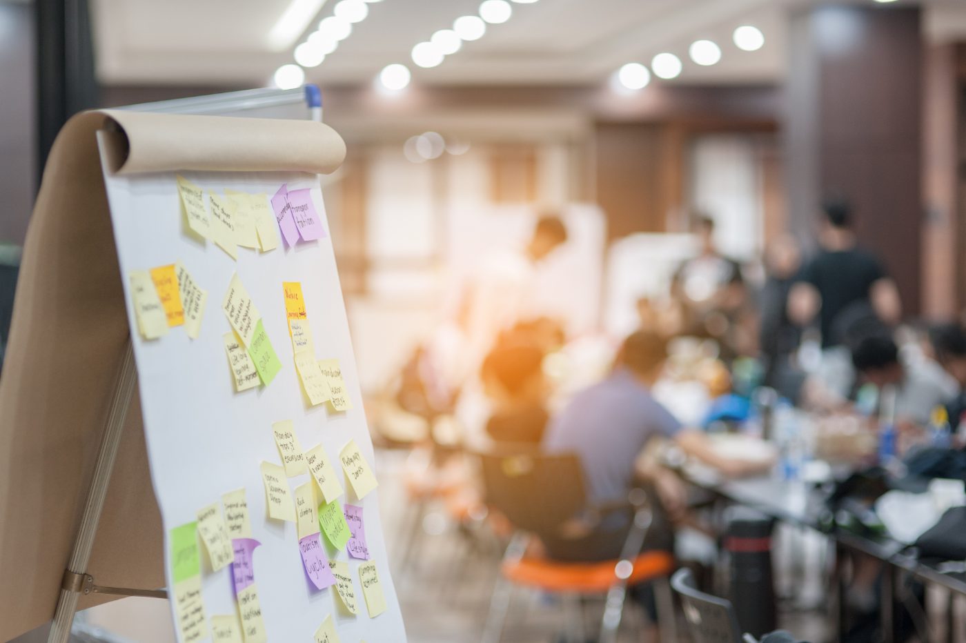 Flipchart mit bunten Haftnotizen, im Hintergrund arbeiten Workshop-Teilnehmende an Tischen